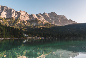 A landscape of Lake Eibsee in Bavaria, southern Germany. The water reflects the distant shore, green forest and mountains of the Alps that climb in the distance. The sky is blue and clear.
