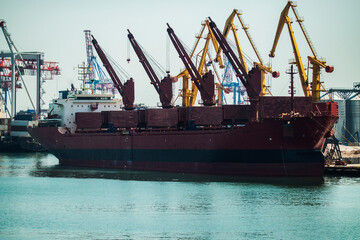 A large red ship stands moored in a seaport against the backdrop of cranes ready for loading