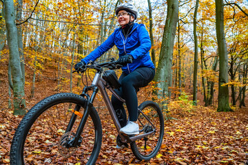 Portrait of middle-aged woman wearing blue sporty jacket, black cycling pants and white bike helmet riding bicycle in forest in autumn scenery. Happy physical activity