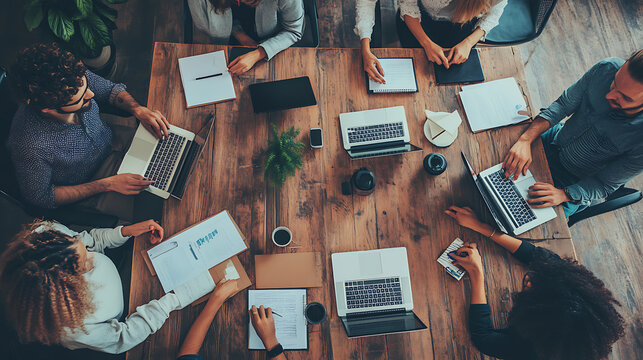 A group of people are sitting around a table with laptops and papers
