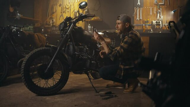 Side view of a confident mechanic in a plaid shirt inspecting motorcycle parts during repairs in his workshop studio
