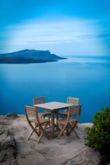Fototapeta premium A wooden table placed on a rocky terrain near the sea. The table has four chairs and is placed near the edge of a cliff. 