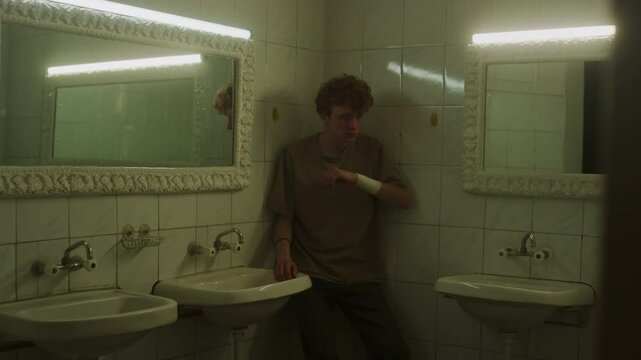 Young man looking at his reflection feeling scared, then hiding in corner between mirrors and sliding down with his back against walls touching face with both hands in public bathroom