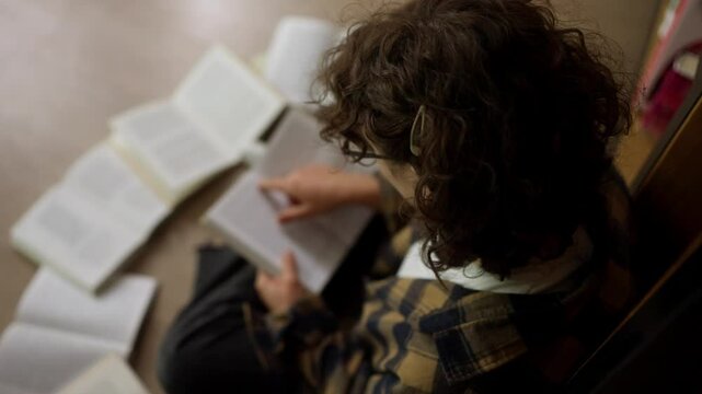 Over the shoulder top view of a student girl with curly hair wearing glasses sitting on the floor and reading a book in the library