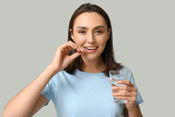 Pretty young woman with glass of water taking pill on grey background