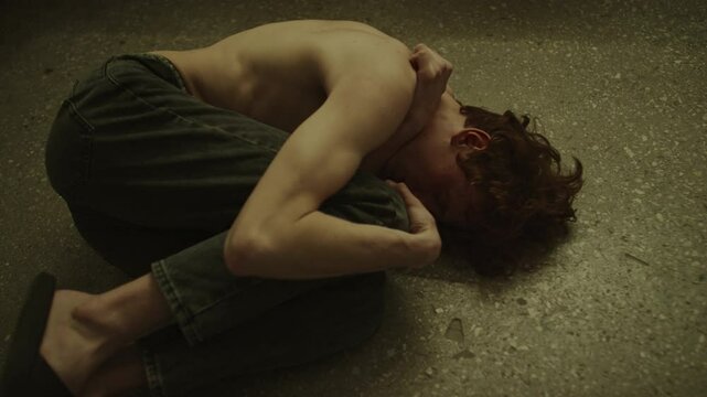 Mentally struggling shirtless young man lying on floor with knees drawn up to chest and arm around knee