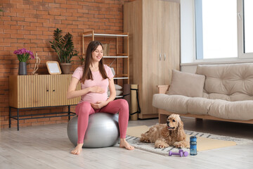 Beautiful sporty young pregnant woman on fitball with cocker spaniel practicing yoga at home