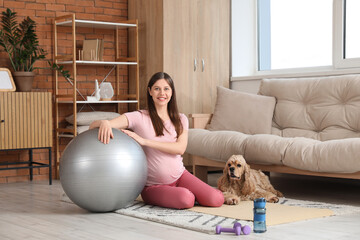 Beautiful sporty young pregnant woman with cocker spaniel and fitball practicing yoga at home