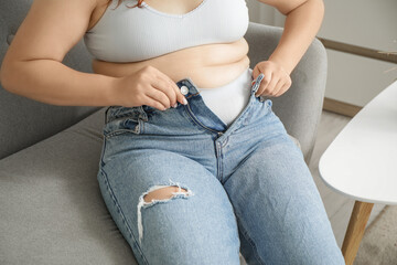 Overweight young woman trying to button tight jeans at home, closeup. Weight gain concept