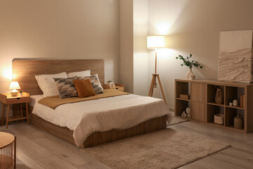 Stylish interior of bedroom with comfortable bed, lamp and shelving unit at evening