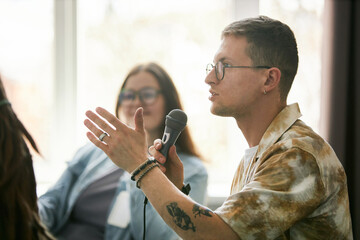 Young man with glasses speaking into microphone during conference event with blurred attendees in background. Wearing casual attire and showcasing engaged facial expression