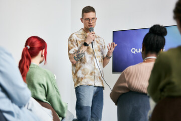 Presenter standing in front of group of diverse people, explaining ideas with microphone and pointing to screen. Audience sitting attentively, focused on presenter