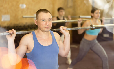 Fototapeta premium Focused middle-aged man in activewear exercising with barbell during group workout in gym health center. CrossFit healthy concept