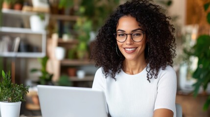 A confident woman with curly hair and glasses is sitting at a desk in an office environment, working on her laptop surrounded by plants, exuding professionalism.