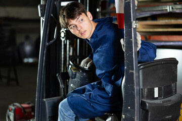 Focused young workman in blue work coat operating lift truck in industrial warehouse setting, looking over shoulder while maneuvering vehicle