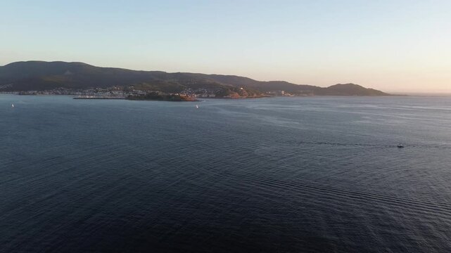 As the golden hour begins, calm ocean waters stretch out, with boats cutting through the sea. In the distance, Baiona appears with its port and parador. The camera descends above Monteferro's coast.