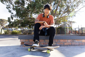 Sitting on skateboard, asian teenage boy with headphones relaxing outdoors in park © wavebreak3