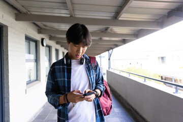 Asian teenage boy using smartphone while standing in school hallway with backpack