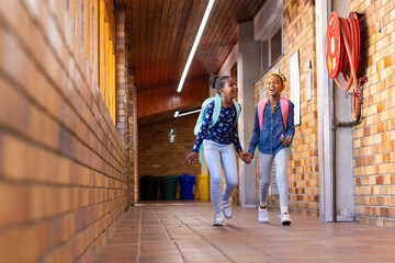 Walking in school hallway, two african american girls with backpacks smiling and holding hands