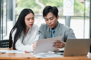 Two young professionals working together on a business project in a modern office environment, reviewing documents and using a laptop.