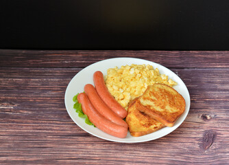 Plate with boiled sausages, egg omelette and bread croutons on a wooden table.