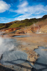 The Krýsuvík/Seltún geothermal area in Iceland features steaming fumaroles, bubbling mud pools, and vibrant sulfuric terrain, set against a backdrop of green hills and a bright blue sky, showcasing th