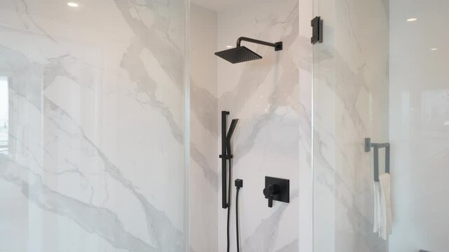 Modern bathroom shower area. Glass door, marble walls with gray veining, black showerhead and hand-held sprayer, white flooring