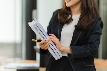 Businesswoman Ready to Present: A professional woman in a sleek black blazer stands confidently, holding a stack of documents and a pen, ready to deliver a compelling presentation. Her focused express