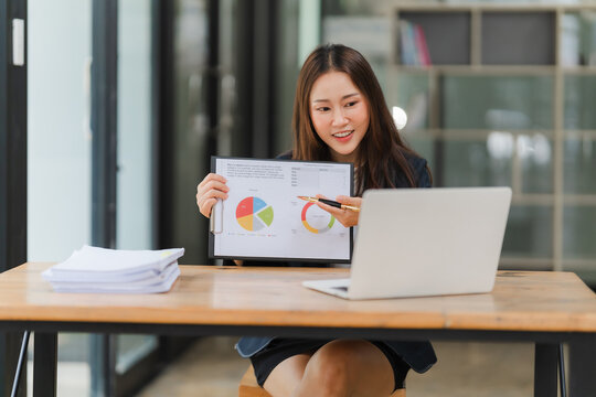 Businesswoman presenting data on video call. A young Asian businesswoman sits at a desk, confidently presenting data on a whiteboard during a video call.  She is using a laptop and a marker to highlig