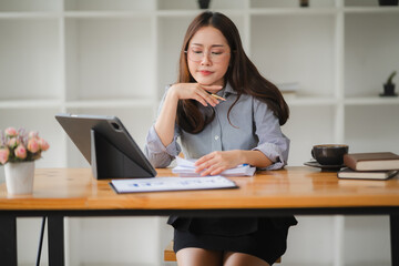 Focused Businesswoman:  A young Asian businesswoman sits at her desk, reviewing documents and data on her tablet, exuding a focused and determined work ethic in a modern office setting.  She is a symb