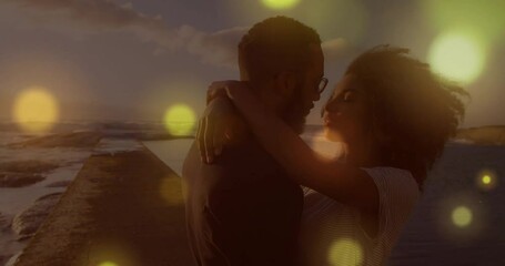 Animation of light spots over biracial couple embracing at seaside - Powered by Adobe