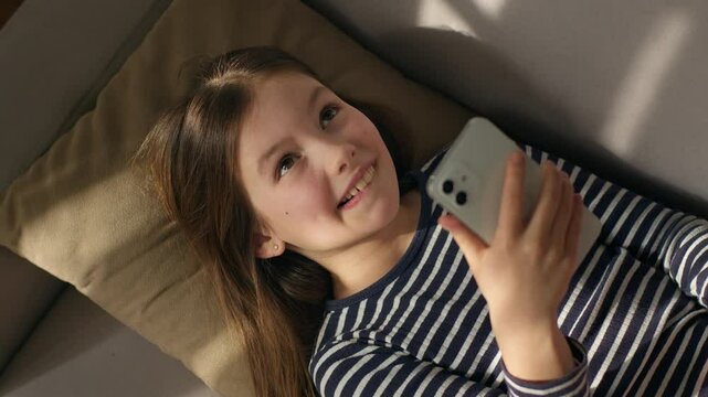 Caucasian European little girl lying on sofa holding using mobile phone smartphone child gadget online texting sms social networks smiling laughing enjoyment watching funny video internet chatting kid
