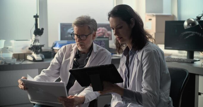 Two diverse scientists in lab coats discuss scientific research, look at papers and use tablet computer. Paleontologists or archaeologists work in archaeological laboratory with modern equipment.