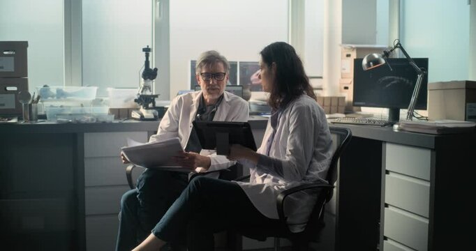 Two diverse scientists in lab coats discuss scientific research, look at papers and use tablet computer. Paleontologists or archaeologists work in archaeological laboratory with modern equipment.