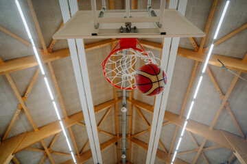 Valmiera, Latvia - April 26, 2024 - A basketball going through the hoop in a gym, captured from below, with the wooden ceiling beams visible in the background.. © Raivo