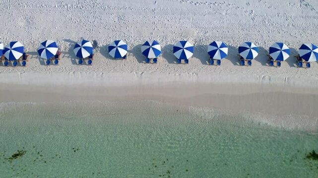 Drone aerial view of blue and white umbrellas on a white sand beach with clear waters on a sunny day