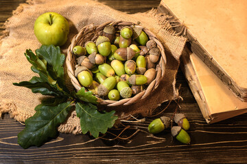wicker basket full of acorns with oak leaves, apple and old books on wooden board, autumn concept