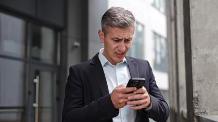 Concerned businessman stands in front of an office building and reads disappointing news on his phone. He is stressed and unsure how to react. Perfect for corporate, finance, and technology themes.