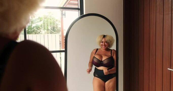 Young African American curvy plus size woman dancing in front of mirror in lingerie and admiring her reflection. Self love and acceptance, positive attitude towards body. Seductive female body.