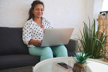 Woman working on a laptop from home on a cozy outdoor patio, surrounded by plants, enjoying a comfortable and productive remote work environment