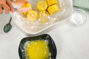 Woman dipping piece of fresh corn in melted butter putting it in baking sleeve on baking sheet on kitchen at home. Prepare bake food dish. Cuisine culinary nutrition domestic food recipe ingredients.