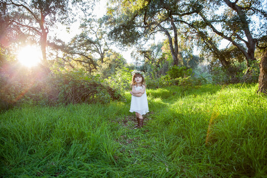 Toddler in a white dress and floral crown standing in a sunlit forest