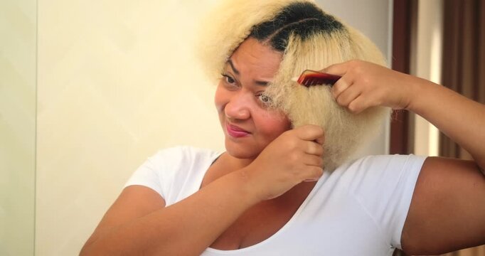 Portrait of a young African American woman standing in front of a mirror trying to comb her curly blonde afro hair. Thick hair is difficult to comb, the comb is stuck. Problem with afro hairstyle.
