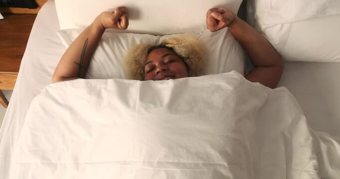 Top view of a young African American woman lying in bed under a white blanket. She is awake and stretching, opening her eyes. Concept of a happy start to the day warm rays of sun illuminating the room