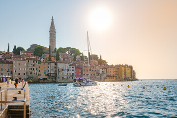 Fototapeta premium The sun casts a warm glow over Rovinj Croatia, illuminating colorful buildings along the waterfront. A sailboat gently floats on the clear waters, inviting exploration of this enchanting coastal town.