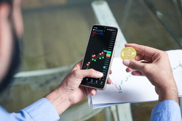 young indian crypto trader or broker using mobile app to check market price for online buying or selling while holding bitcoin in hand. Investment. Finance and economy. Digital money or asset. 