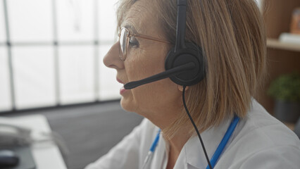 Elderly, blonde, woman with glasses and headset working in a clinic, focused on a conversation, captures the essence of professional medical support in a modern indoor setting.