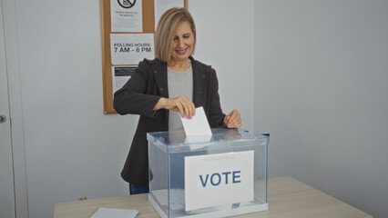 Elderly blonde woman voting in an indoor electoral college room, placing her ballot in a...