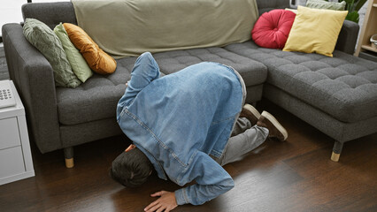 Young man in denim searching under couch in modern living room interior