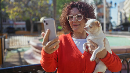 Elderly woman holding a chihuahua while taking a selfie with a smartphone in an urban park.